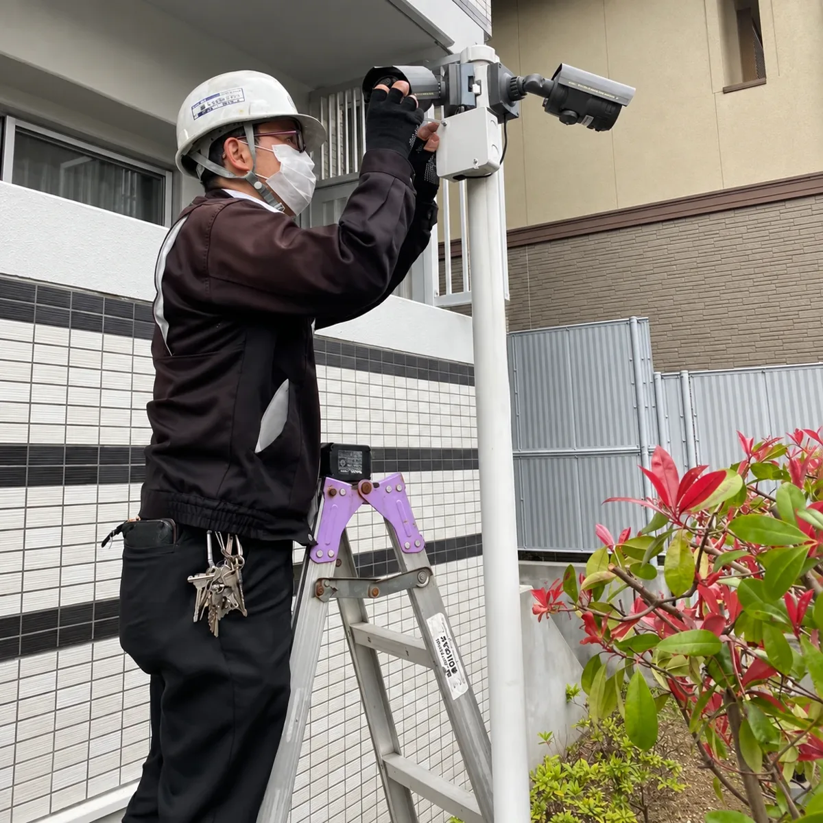 脚立に上り、建物の外壁沿いのポールに防犯カメラを取り付ける当社スタッフ
